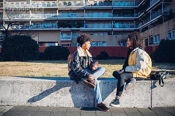 Zwei coole junge Freundinnen sitzen auf der Stadtmauer und unterhalten sich