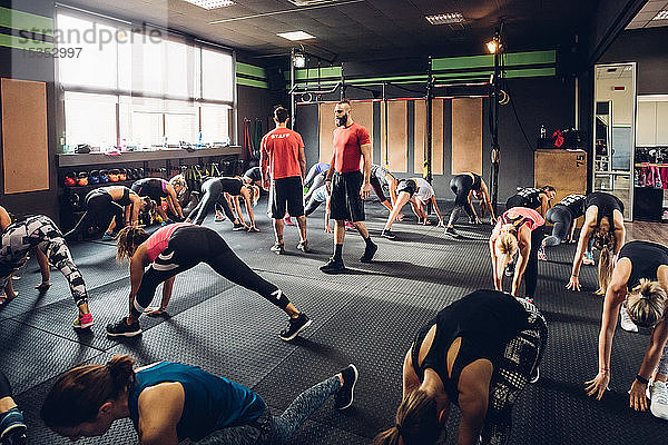 Große Gruppe von Frauen trainiert in Turnhalle mit männlichen Trainern  beugt sich mit ausgestreckten Beinen vor