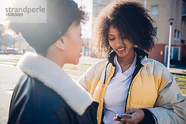 Zwei coole junge Freundinnen schauen sich ein Smartphone in einer städtischen Straße an