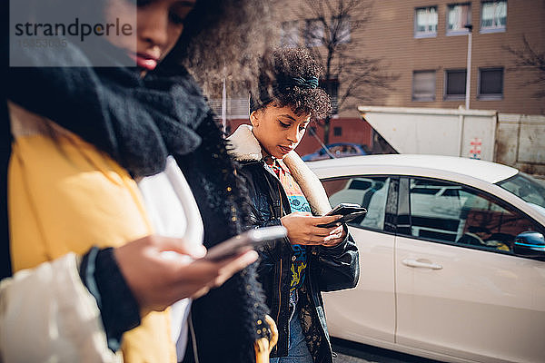 Zwei coole junge Frauen  die auf dem städtischen Bürgersteig spazieren gehen und auf Smartphones schauen  Seitenansicht