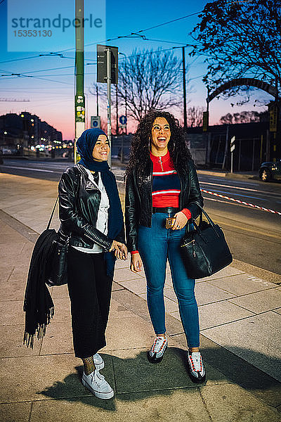 Junge Frau im Hidschab mit bester Freundin auf dem Bürgersteig der Stadt in der Dämmerung  in voller Länge