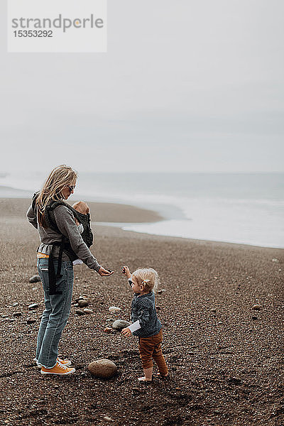 Mutter mit Baby in Tragetasche spielt mit Kleinkind am Strand