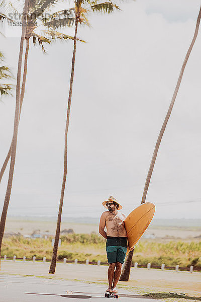 Mittelgroßer erwachsener männlicher Skateboarder mit Surfbrett  stehend auf einem Skateboard auf der Küstenstraße  Haiku  Hawaii  USA