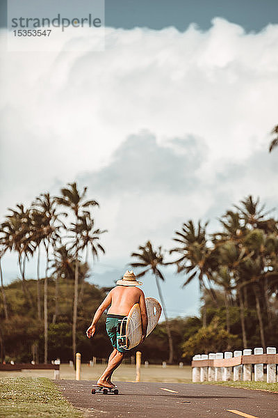 Mittelgroßer erwachsener männlicher Skateboarder mit Surfbrett  Skateboarding auf Landstraße  Rückansicht  Haiku  Hawaii  USA