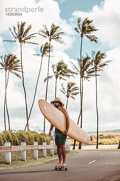 Mittelgroßer erwachsener männlicher Skateboarder mit Surfbrett  Skateboarding auf Küstenstraße  Haiku  Hawaii  USA