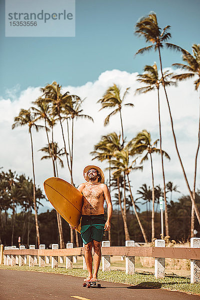 Mittelgroßer erwachsener männlicher Skateboarder mit Strohhut  steht auf Skateboard und trägt ein Surfbrett auf einer Landstraße  Portrait  Haiku  Hawaii  USA