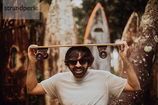 Mittelgroßer erwachsener männlicher Skateboarder  der ein Skateboard auf dem Kopf trägt  nach alter Surfbrettstruktur im Park  Portrait  Haiku  Hawaii  USA