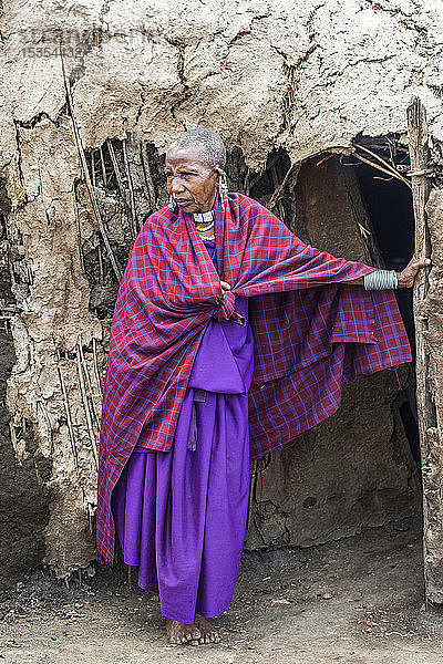 Älteste Maasai-Frau in traditioneller Kleidung steht am Eingang ihrer Lehmhütte im Ngorongoro-Schutzgebiet; Tansania