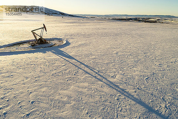 Luftaufnahme von Pumpjack in einem schneebedeckten Feld mit langen Schatten  warmem Licht des Sonnenaufgangs  sanften Hügeln  schneebedeckten Bergen und blauem Himmel im Hintergrund; Longview  Alberta  Kanada