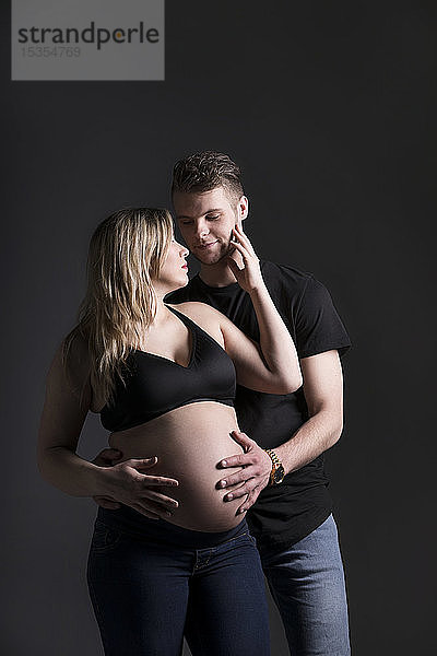 Ein junges  schwangeres Paar schaut sich an  und der Vater hält ihren Bauch in einem Studio vor einem dunklen Hintergrund: Edmonton  Alberta  Kanada