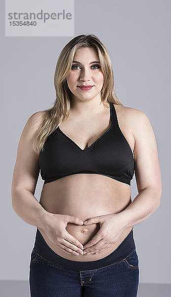 Eine junge schwangere Frau hält ihren Bauch in einem Studio und posiert für die Kamera  während sie ein Herz über ihrem ungeborenen Kind formt; Edmonton  Alberta  Kanada