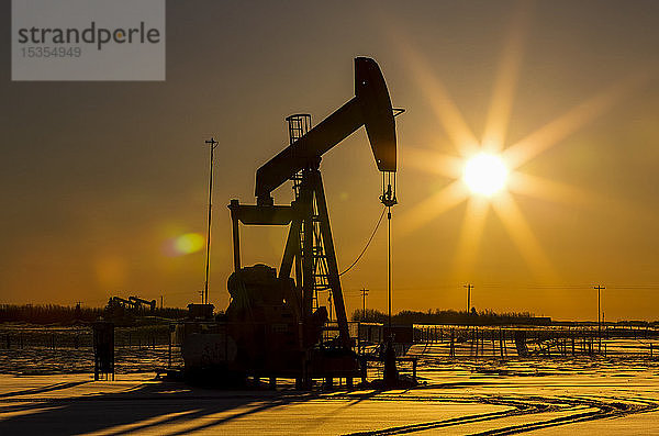 Silhouette eines Pumpjacks in einem schneebedeckten Gebiet mit einer orangefarbenen Starburst-Sonne bei Sonnenaufgang; Longview  Alberta  Kanada