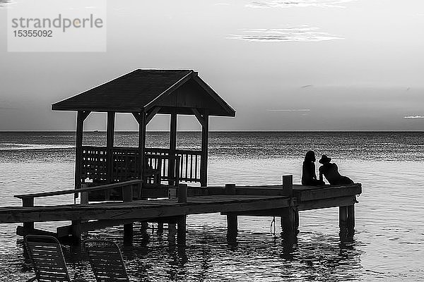 Ein Paar sitzt am Ende eines Stegs und blickt bei Sonnenuntergang auf den ruhigen Ozean; Roatan  Bay Islands Department  Honduras