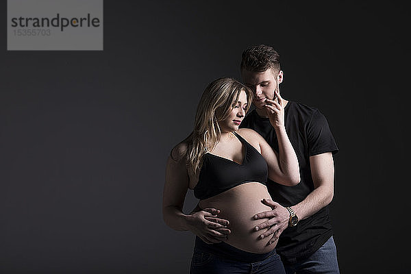 Ein junges schwangeres Paar  das auf sein Baby herabblickt  während der Vater ihren Bauch in einem Studio vor einem dunklen Hintergrund hält: Edmonton  Alberta  Kanada