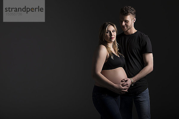 Ein junges schwangeres Paar mit der Mutter  die in die Kamera schaut  während sie ihren Bauch in einem Studio vor einem schwarzen Hintergrund hält: Edmonton  Alberta  Kanada
