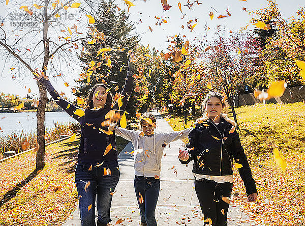 Eine Mutter und ihre beiden Töchter gehen in einem Stadtpark an einem warmen Herbsttag spazieren und werfen Blätter in die Luft; Edmonton  Alberta  Kanada