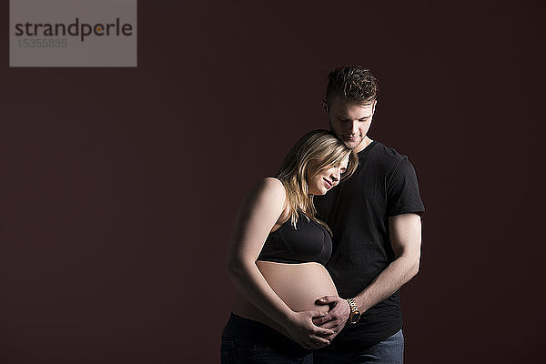 Ein junges schwangeres Paar  das auf sein Baby herabblickt  während es seinen Bauch in einem Studio vor einem schwarzen Hintergrund hält: Edmonton  Alberta  Kanada