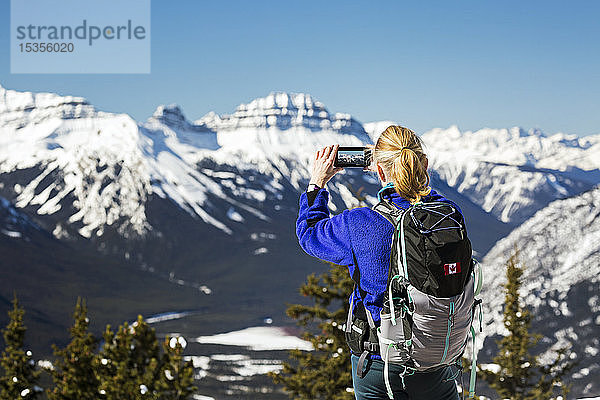 Wanderin auf dem Gipfel eines Berges  die mit ihrem Handy ein Foto von einer schneebedeckten Bergkette mit blauem Himmel macht; Banff  Alberta  Kanada