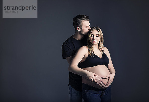 Ein junges schwangeres Paar sieht sich an  und der Vater küsst ihre Stirn und hält ihren Bauch in einem Studio vor einem dunklen Hintergrund; Edmonton  Alberta  Kanada