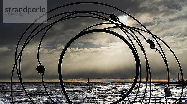 Silhouette einer Skulptur und des Roker-Leuchtturms in der Ferne entlang der Uferlinie des Flusses Ware mit Gewitterwolken über dem Kopf; Sunderland  Tyne and Wear  England