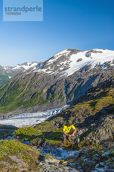 Ein Mann  der mit dem Rucksack unterwegs ist und seine Wasserflasche an einem kleinen Bach im Kenai Fjords National Park mit dem Exit Glacier im Hintergrund an einem sonnigen Sommertag in Süd-Zentral-Alaska auffüllt; Alaska  Vereinigte Staaten von Amerika