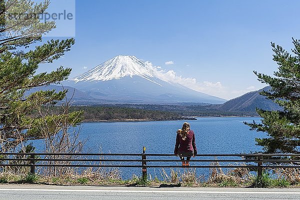 Junge Frau sitzt auf einem Geländer neben einer Straße und blickt über den See auf den Vulkan Mt. Fuji  Motosu-See  Präfektur Yamanashi  Japan  Asien