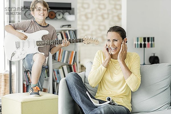 Mutter sitzt lachend auf der Couch im Wohnzimmer und hält sich die Ohren zu  Sohn spielt E-Gitarre  Deutschland  Europa