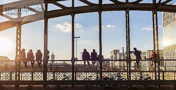 Junge Leute sitzen auf dem Geländer der Hackerbrücke über die Bahngleise und schauen in die Ferne  Gegenlicht  München  Oberbayern  Bayern  Deutschland  Europa