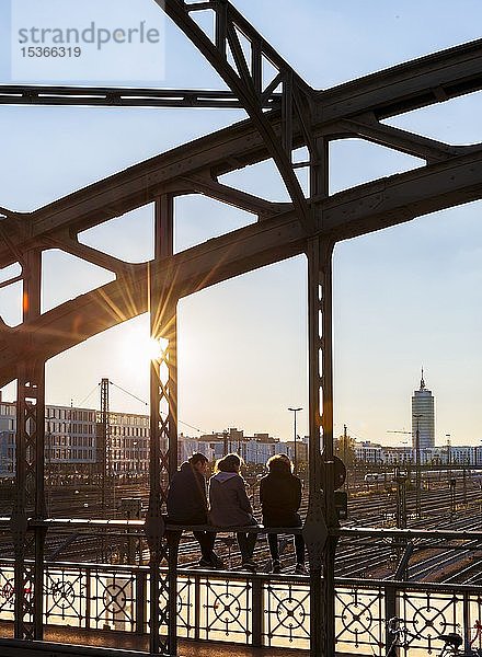 Drei Jugendliche sitzen auf der Brüstung der Hackerbrücke über die Bahngleise und schauen in die Ferne  Gegenlicht  München  Oberbayern  Bayern  Deutschland  Europa
