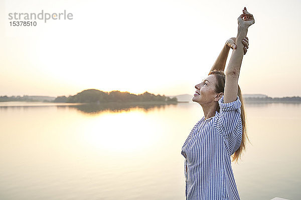 Glückliche reife Frau streckt sich bei Sonnenaufgang an einem See