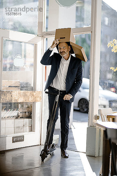 Geschäftsmann mit Pappkarton auf dem Kopf  auf einem E-Scooter in einem Café