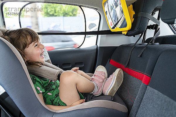 Lachendes Kleinkind Mädchen sitzt auf einem Autositz und schaut in einen Spiegel