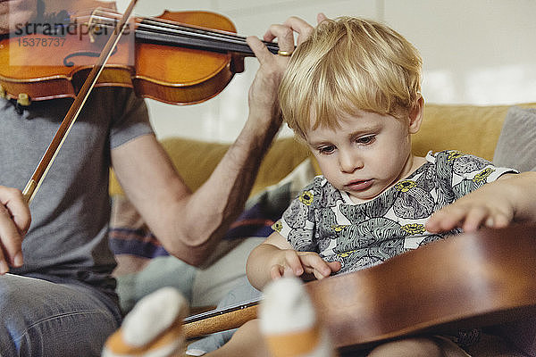Porträt eines Kleinkindes beim Ukulele-Test  während sein Vater Geige spielt