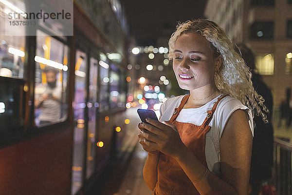 Junge Frau in London schaut nachts auf ihr Smartphone  im Hintergrund der Bus
