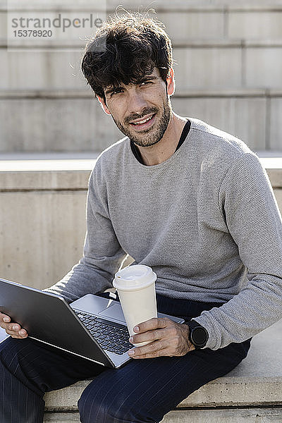 Mann sitzt auf einer Außentreppe mit Kaffee zum Mitnehmen und Laptop