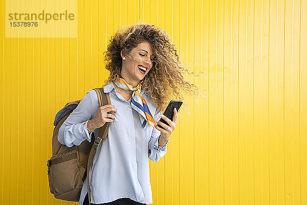 Lachende junge Frau mit Rucksack vor gelbem Hintergrund schaut auf Handy