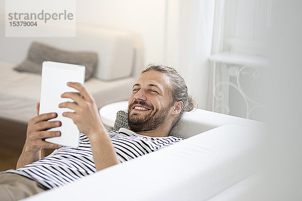 Lächelnder junger Mann liegt zu Hause auf Couch und benutzt Tablette