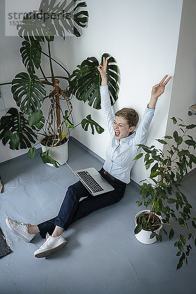 Jubelnde Geschäftsfrau mit Laptop sitzt auf dem Boden  umgeben von Pflanzen