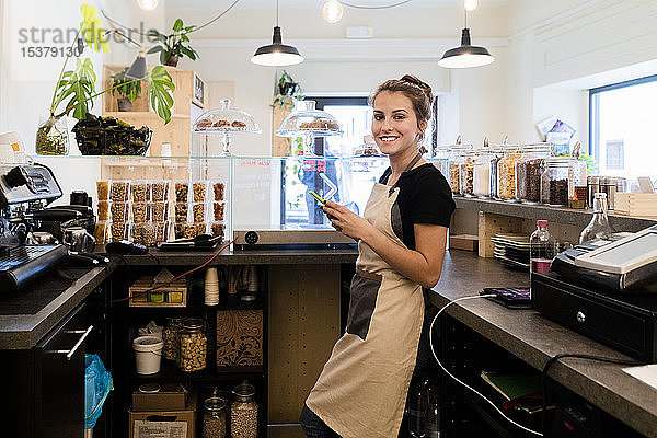 Porträt einer lächelnden jungen Frau mit Handy hinter dem Tresen in einem Cafe