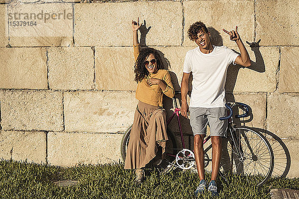 Junges Paar mit Fahrrad  an Steinmauer gelehnt  Handzeichen machen
