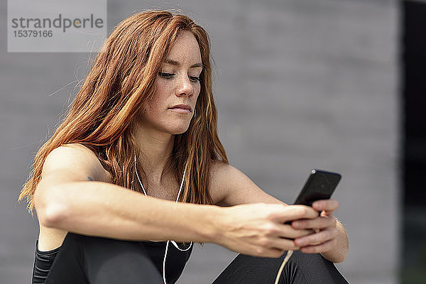 Sportliche junge Frau mit Kopfhörern macht eine Pause und benutzt das Smartphone im Freien