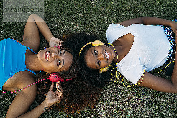 Porträt von zwei glücklichen jungen Frauen  die auf einer Wiese liegen und mit Kopfhörern Musik hören