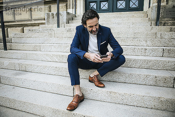 Bärtiger  reifer Geschäftsmann in blauem Anzug sitzt auf der Treppe und schaut auf sein Handy