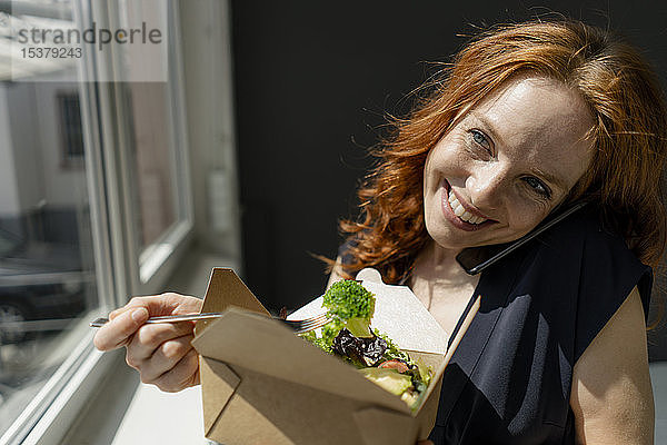 Porträt einer fröhlichen rothaarigen Geschäftsfrau  die gesundes Essen zum Mitnehmen isst und telefoniert