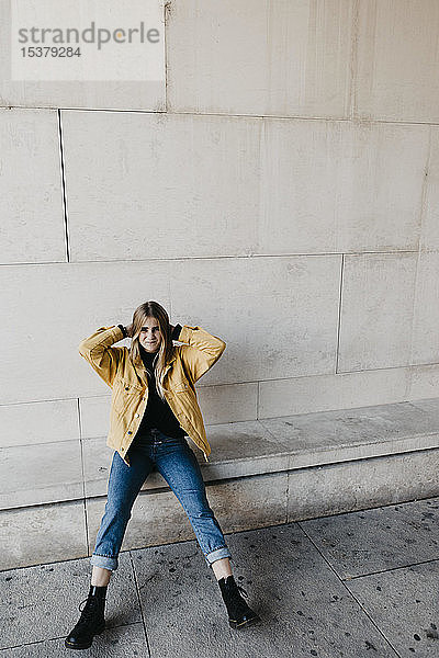 Porträt einer jungen Frau in Jeans und gelber Jacke  die an einer Wand sitzt