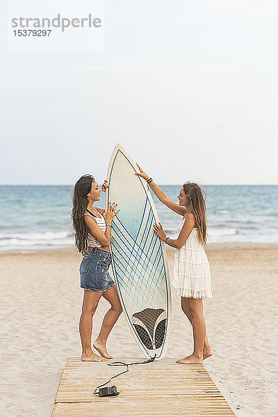 Zwei glückliche Freundinnen mit Surfbrett auf der Strandpromenade stehend