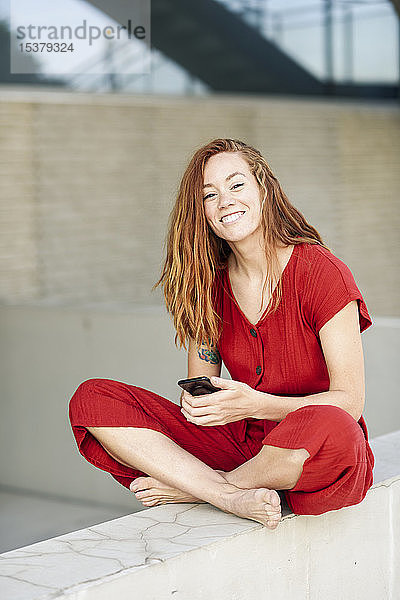 Porträt einer lächelnden  sportlichen jungen Frau  die eine Pause macht und ihr Smartphone im Freien benutzt