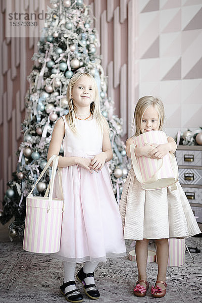 Porträt von zwei glücklichen kleinen Mädchen mit Geschenkkartons vor beleuchtetem Weihnachtsbaum stehend