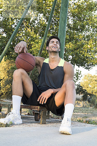 Junger Sportler sitzt auf dem Sportplatz  hält Basketball in der Hand und lacht  während er wegschaut