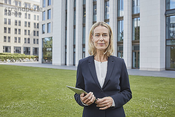 Porträt einer selbstbewussten Geschäftsfrau mit Tablette auf dem Rasen in der Stadt stehend